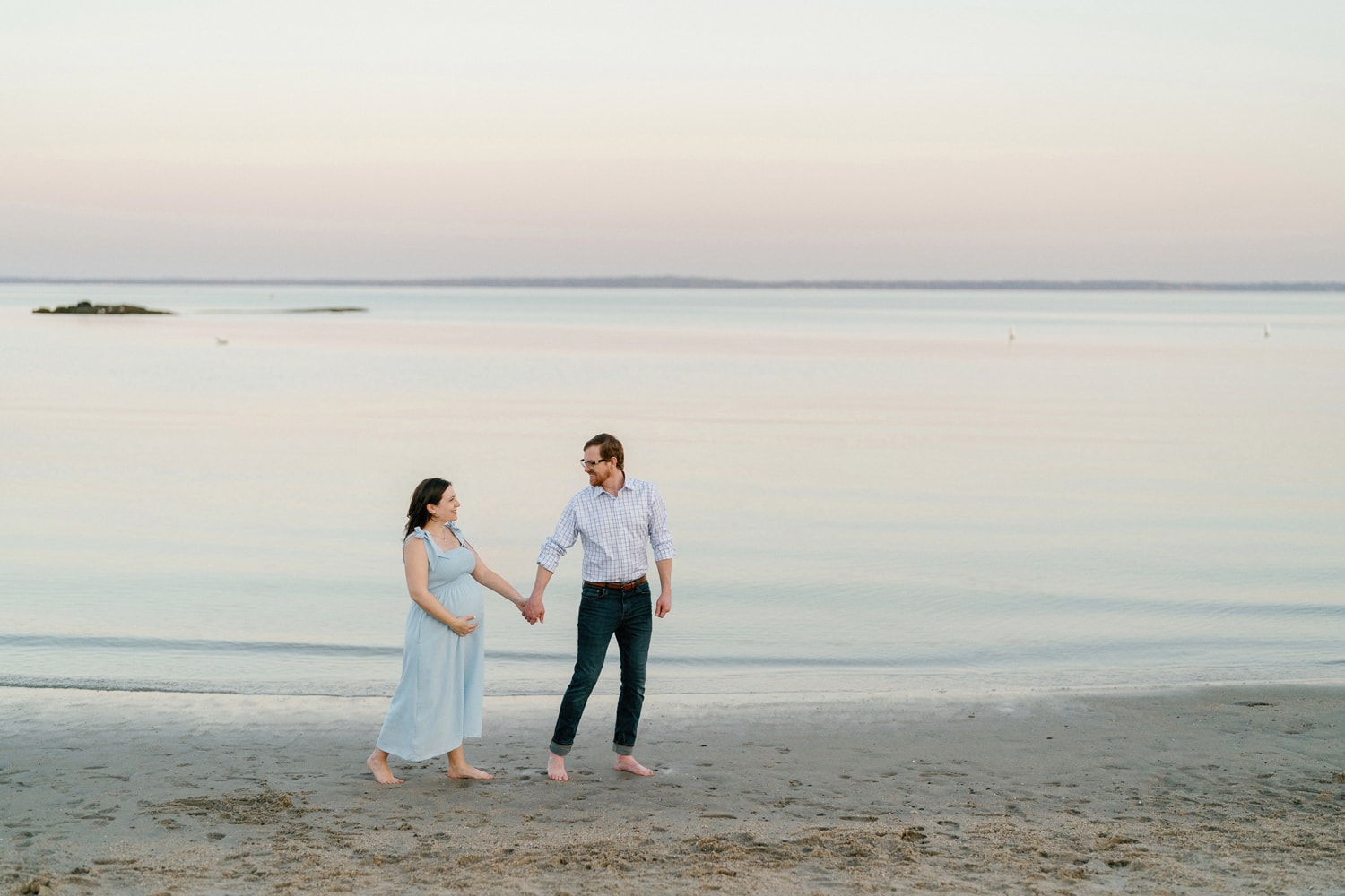 A couple in Westchester, NY, strolling hand in hand near the water on the beach during a maternity photoshoot.