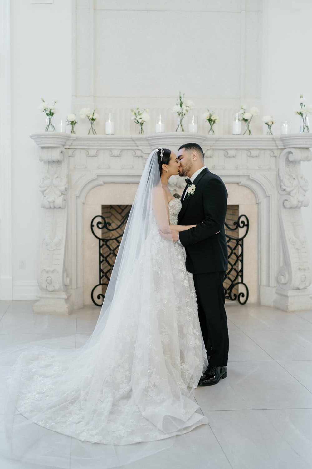 A bride and groom share a romantic kiss during their indoor wedding ceremony in The Great Hall at The Estate at Florentine Gardens, standing in front of an elegant white fireplace adorned with candles and floral arrangements.