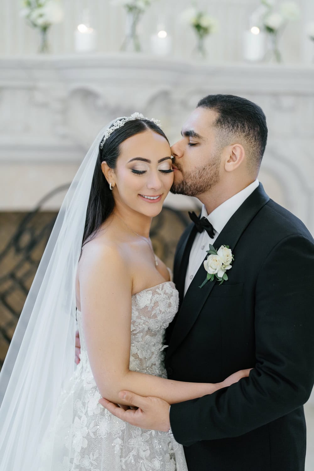 A groom gently kisses his bride’s forehead during their intimate indoor wedding ceremony at The Estate at Florentine Gardens, with a beautifully decorated fireplace in the background.