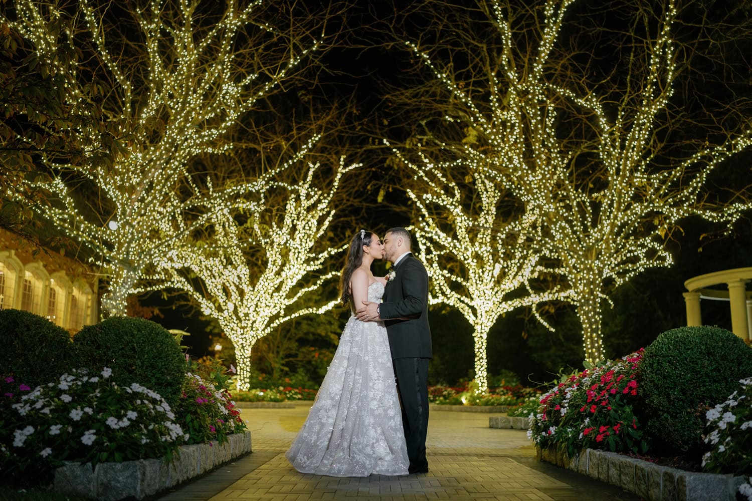 A bride and groom share a romantic kiss in the illuminated gardens of The Estate at Florentine Gardens at night, surrounded by twinkling fairy lights and lush floral landscaping.