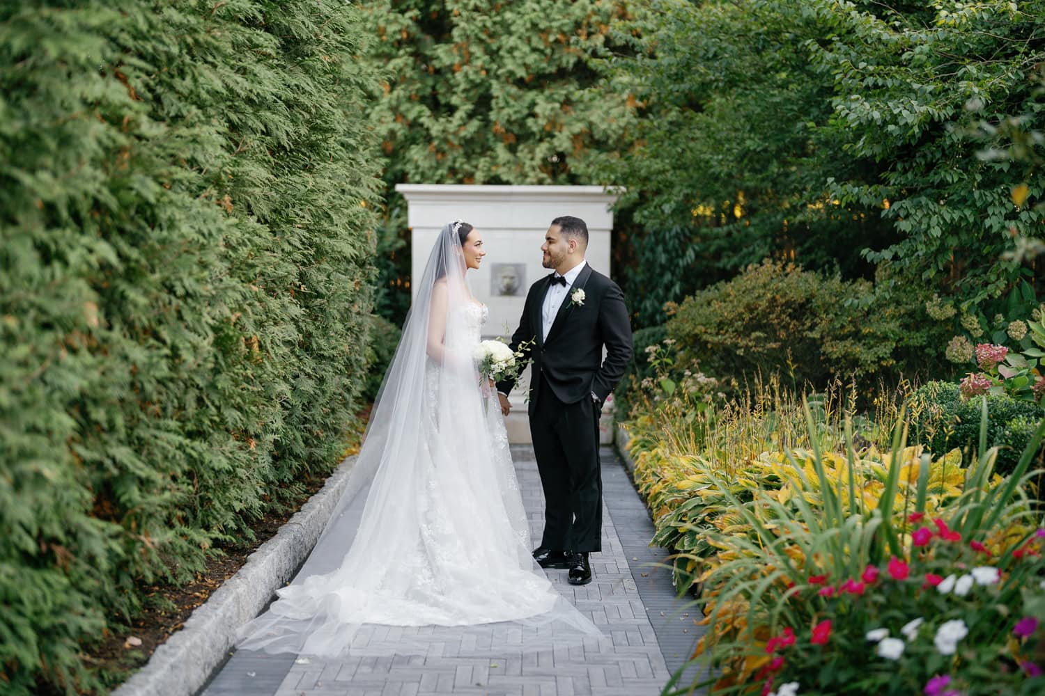 A bride and groom share a quiet moment on a picturesque garden pathway at The Estate at Florentine Gardens, surrounded by lush greenery and vibrant florals, creating a perfect backdrop for timeless wedding portraits.