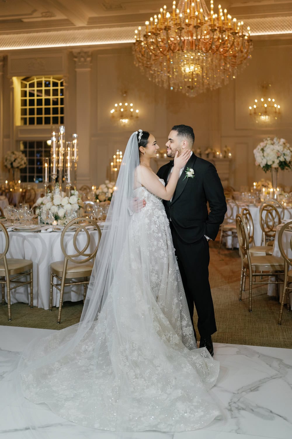 A newlywed couple embraces under the grand chandelier in the Florentine Gardens ballroom, with candlelit tables and floral arrangements setting the perfect romantic reception ambiance.