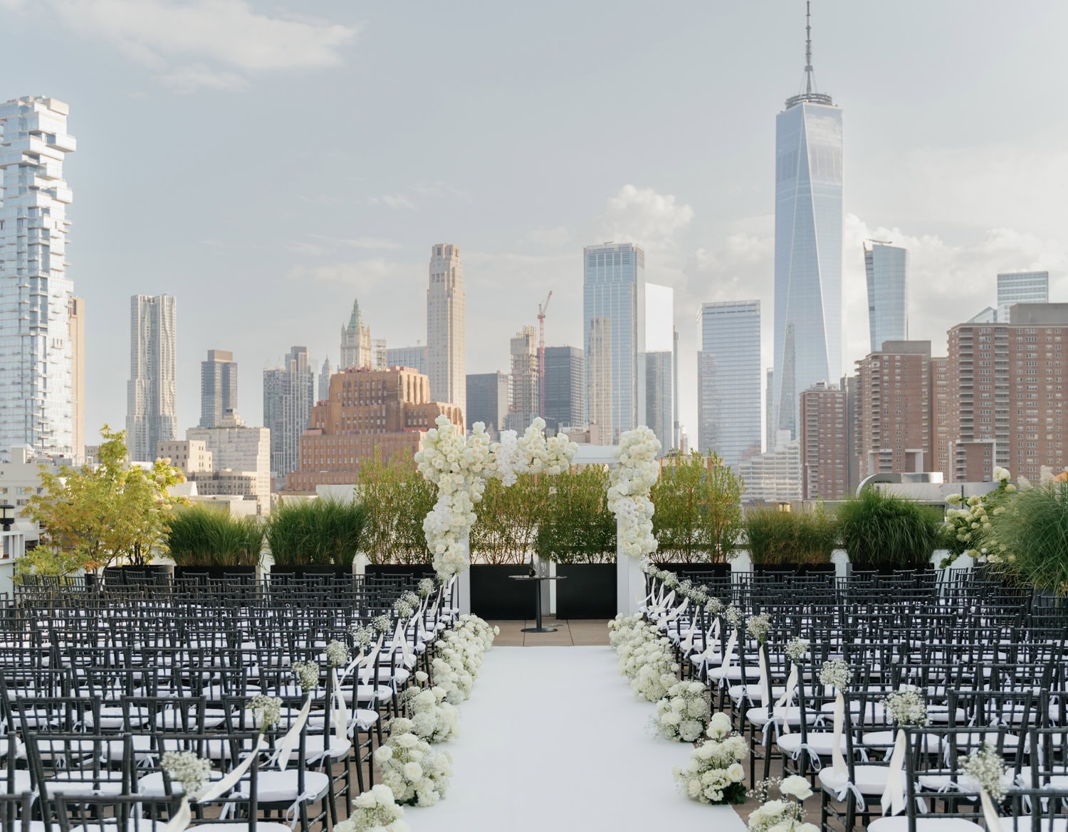 A stunning rooftop wedding ceremony at Tribeca Rooftop in NYC with a breathtaking skyline view, elegant floral arrangements, and modern black Chiavari chairs.