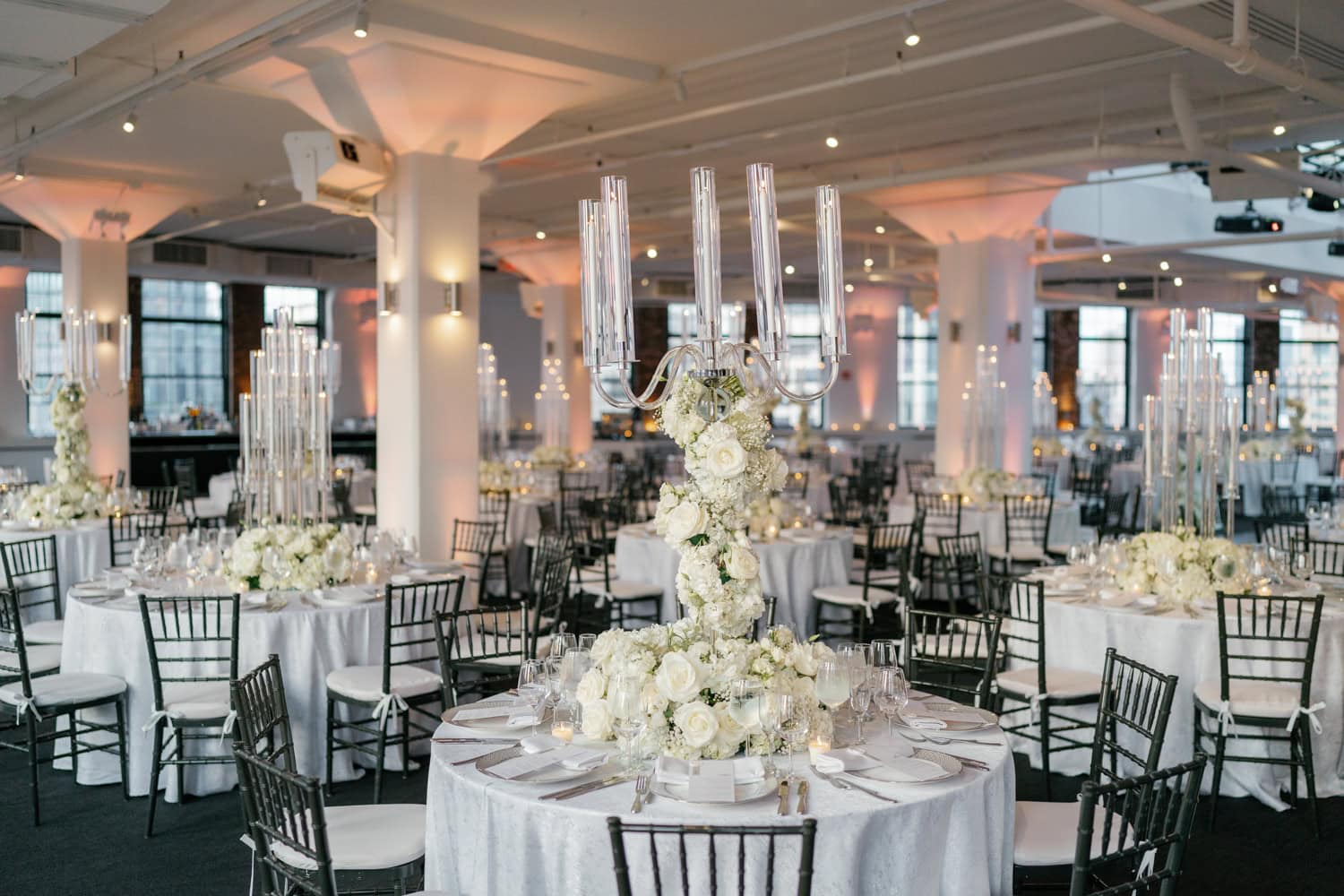 Elegant wedding reception setup, featuring luxurious floral centerpieces, tall candle holders, and chic black Chiavari chairs in a modern loft-style venue.