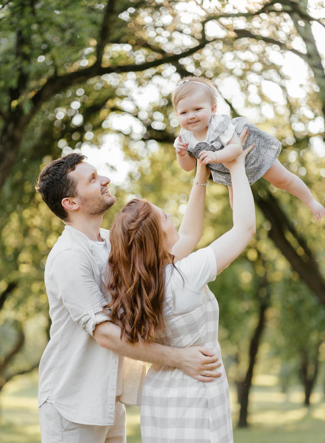 Parents lifting their baby in the air during a sunlit outdoor family session, capturing a candid and playful moment.