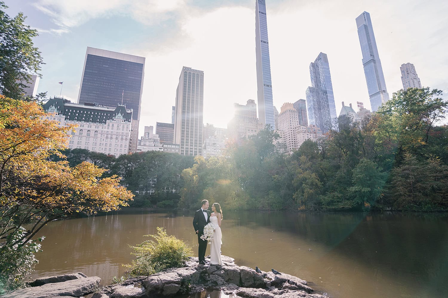 Bride and groom standing on a rock in Central Park with the NYC skyline in the background, capturing a romantic wedding moment near Essex House.