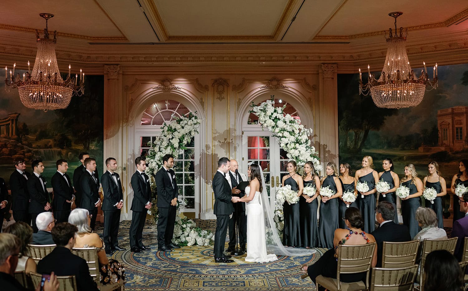 A couple exchanges vows during an elegant indoor wedding ceremony at JW Marriott Essex House, surrounded by a bridal party in black attire and floral arch décor.