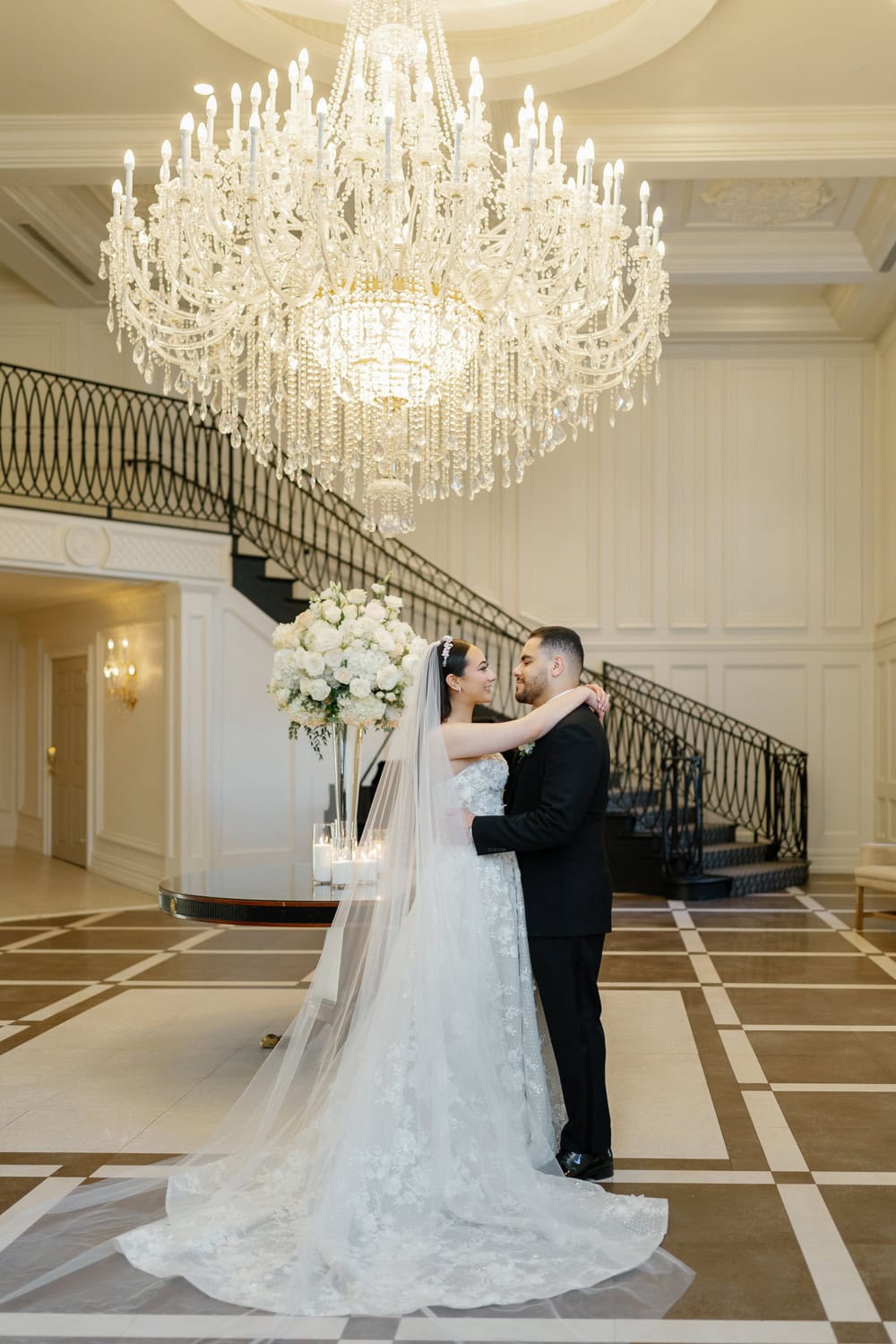 A newlywed couple shares a romantic moment beneath the grand crystal chandelier in the foyer of The Estate at Florentine Gardens, highlighting the venue’s sophisticated and elegant design.