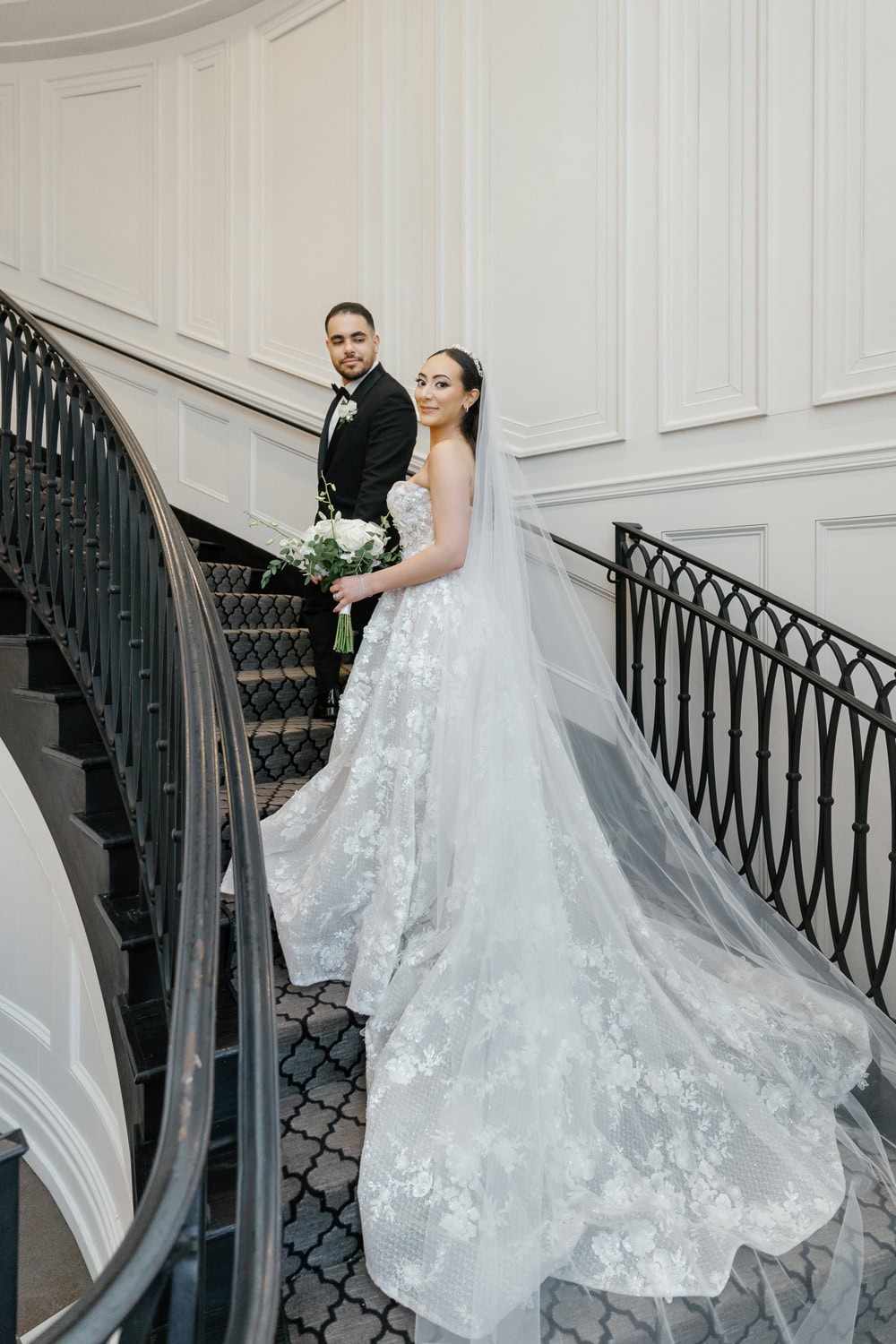 A bride and groom pose on the elegant wrought-iron staircase in the grand foyer of The Estate at Florentine Gardens, showcasing the venue’s classic architectural beauty.