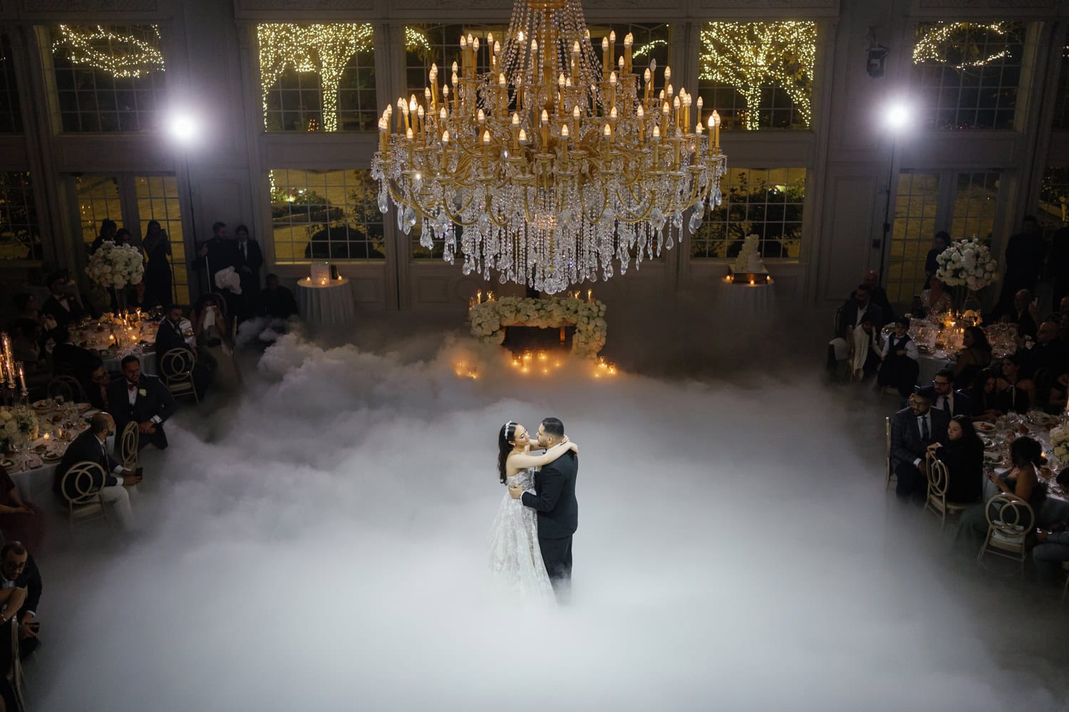 A bride and groom share their romantic first dance at The Estate at Florentine Gardens, surrounded by a dreamy cloud effect under a grand crystal chandelier in the ballroom, with guests watching from elegantly set tables.