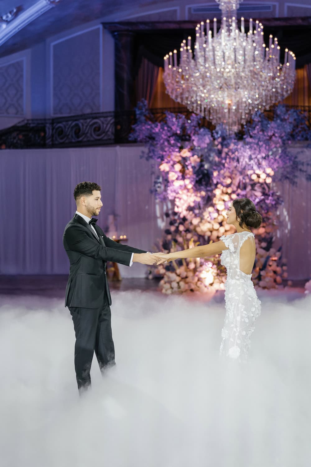 A bride and groom share a romantic first dance, surrounded by elegant floral arrangements and a grand chandelier, with a dreamy fog effect enhancing the atmosphere