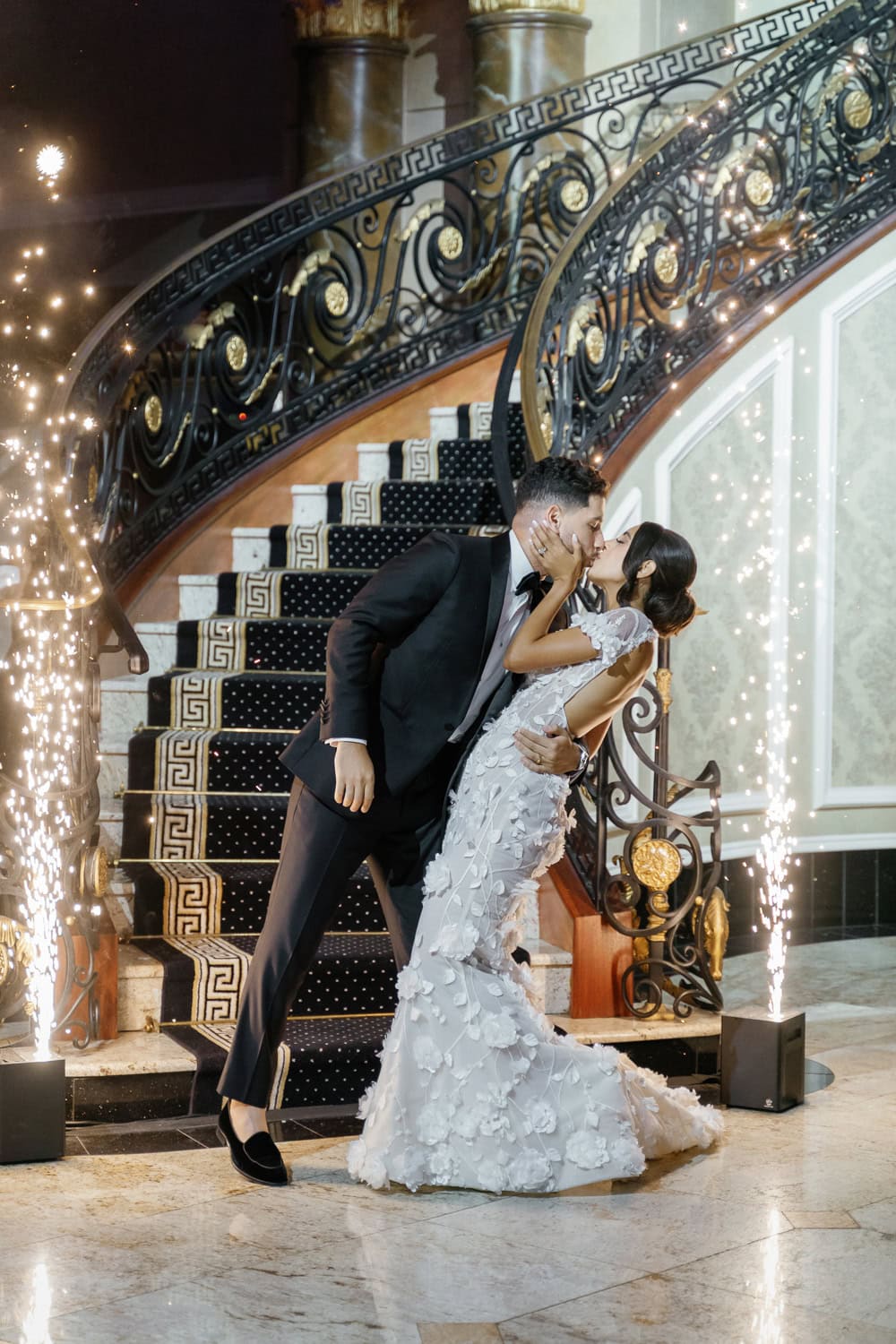 A newlywed couple shares a romantic kiss at the base of the grand staircase in the Palazzo Ballroom, with sparklers creating a magical entrance.