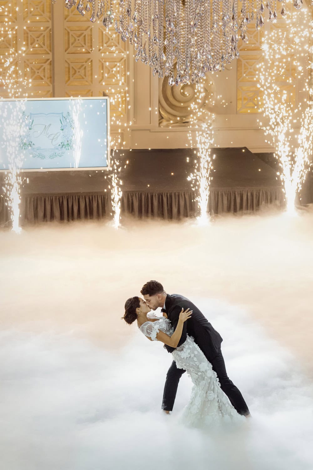 A newlywed couple shares a passionate dip and kiss during their wedding reception, with sparklers and a cloud-like dance floor creating a breathtaking moment.