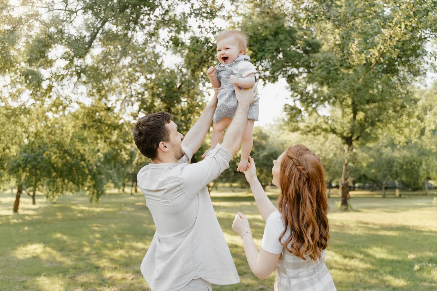 Father lifting smiling baby in the air while mother watches during a family photography session in Ardsley, NY, surrounded by greenery.